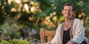 Professor Sally Otto seated on a wooden bench in a garden, smiling gently. She has short brown/black hair, round glasses, and is wearing a cream-coloured cardigan over a black loose blouse.